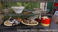 Two shelves holding assorted glassware, stoneware, collectible items, an iron, and serving platters arranged together for sale. The shelving is metal and not included in the lot.