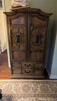 Front view of vintage solid wood armoire showing carved wooden doors with geometric patterns and three drawers below. The armoire is approximately 6 feet tall and made of heavy, solid wood with some signs of wear.