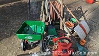 Electric leaf blower, green push spreader, garden rake, coiled wires, garden hoses, terracotta pots, and snow shovel shown outdoors on pavement