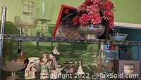 Two shelves of collectibles featuring artificial flower arrangements, antique ceramic figurines, depression glass, glassware items, and Christmas ware.