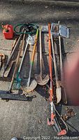 Overview of gardening tools and gas can arranged on pavement, includes shovels, rake, hose and orange handled hand tool