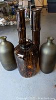 Five decorative colored glass vases on floor. Two tall dark brown bamboo-style vases in back, two olive green vases on sides, one round mottled dark brown vase front center.