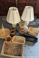 Photo showing the pair of off-white lamps with matching pleated shades, a collection of assorted woven baskets of various sizes and styles including Native American made baskets, and a glass potpourri container with dried flowers inside.