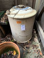 Full view of large round stoneware crock with lid placed outdoors on ground, dirty with leaves and debris