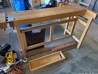 Full side view of beech workbench on concrete floor showing dog hole grid on top, leg framework and detached wooden tray underneath.