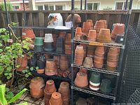 Left and right metal wire shelving racks holding various terra-cotta and plastic pots outdoors against wooden fence.