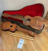 Two ukuleles and drum sticks arranged with open red-velour-lined hard case