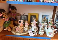 Wide view showing the two stuffed Hawaiian dolls, various ceramic figurines including children and cherubs, ceramic pitcher and trinket boxes on a wooden shelf.