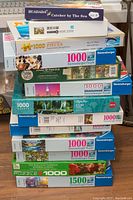 Stack of 13 assorted jigsaw puzzle boxes, mainly Ravensburger brand, showing various puzzle images and piece counts including 1000 and 1500 pieces