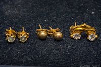 Photo showing all three pairs of gold stud earrings on a dark fabric background.