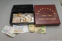 Black collector's box filled with assorted coins and several currency notes placed outside, next to a wooden box labeled in gold Chinese characters 1949-1999 commemorative coin set.