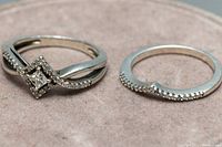 Pair of sterling silver rings displayed on a suede-like pink surface, showing top and side views of diamond settings and ring bands.