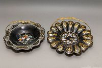 Two black paper-mache cake baskets with scalloped edges, brass handles, gold painted decoration, and mother of pearl inlays, placed side by side on a beige surface.