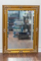 Full view of large rectangular wall mirror with ornate gold frame and beveled glass edges leaning against a white wall.