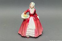 Front left angle view of the porcelain figurine showing the red dress with white apron and flowers in basket.