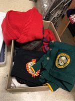 Box of various unisex sweaters including red, green, black with patches and a patterned one.