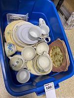 Top view of blue bin containing assorted vintage china including cups, saucers, plates, and serving dishes mostly in ivory and cream colors with floral decorations.