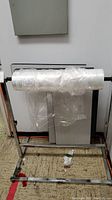 Front view of metal garment bag stand holding clear plastic wrap roll against a wall.