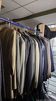 Photo showing multiple men's blazers hanging on a blue garment rack, mostly in neutral colors including beige, grey, and patterned fabrics.