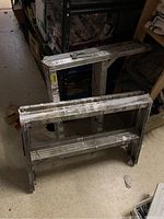 Two aluminum saw horses standing next to each other in storage, showing dimensions and worn condition.