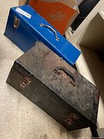 Two metal tool boxes on floor, one blue and one black with diamond plate pattern, both with handles and latches.