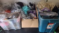 Three storage containers filled with assorted wigs in plastic bags, including curly blondes, black, and turquoise wigs