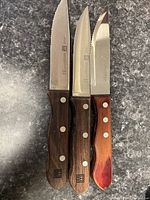 Photo of three serrated steak knives on a gray countertop, showing handles and blades. Two are Zwilling J.A. Henckels with wooden handles. One is a Paderno with a red-stained handle.