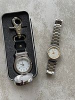 Photo showing both watches from front view: one clip-on style watch with white dial and black numerals on nylon strap with metal clip; one metal bracelet wristwatch with white dial, gold hands and bezel.