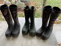Front view of three pairs of hunting boots on a table outside showing two pairs with camouflage on sides and one solid green pair in middle.