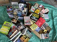 Five boxes of miscellaneous books arranged on a tarp. Covers of various books including Janet Evanovich, John Grisham, Robert Crais, and Ngaio Marsh are visible.