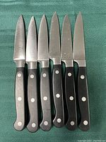 Photo showing six Chicago Cutlery paring knives with stainless steel blades and black riveted handles arranged on a green striped textured surface.