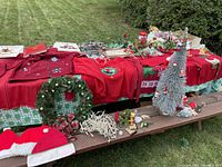Overview of lot with Christmas sweaters, wreath, light string, bows, floral picks, Santa hats, and fiber optic tree displayed on table and bench.