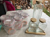 Six Luminarc glass canning jars with clamps and rubber seals placed together on a table, alongside three glass jars with wooden lids on a wooden base. One glass jar missing from original set of four in wooden lid group.