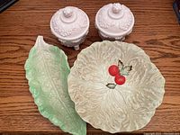 Four ceramic pieces on wooden table including two white lidded jars, a green leaf-shaped plate, and a large cabbage-leaf style bowl with cherry decoration