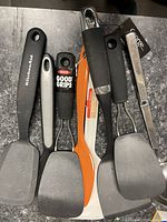 Top view of 7 kitchen utensils including spatulas, ladle, and masher showing handles and utensil heads side by side on dark countertop.
