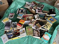 Multiple cardboard boxes filled with various used books placed on a green tarp, showing a diverse collection mostly paperback with different sizes and genres.