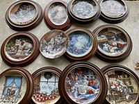Photo showing all 12 round framed cat collector plates arranged on a floor, depicting various indoor and outdoor feline scenes with wood frames, some with minor damage.