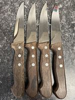 Set of four steak knives with wooden handles and serrated blades lying flat on dark countertop showing overall shape and condition.