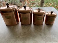 Four vintage copper canisters in a line outdoors, labeled Flour, Sugar, Coffee, and Tea. All have lids with decorative flower-like knobs.