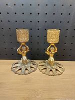 Two decorative metal candle holders side by side on wooden surface with black pegboard backdrop, showing front and side angles