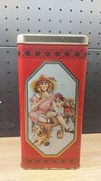 Front side of tin with illustration of a young girl sitting with a doll and teddy bears on a red background.