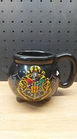 Front and side views of black ceramic Harry Potter mug decorated with colorful Hogwarts crest and Latin motto. The mug has a round body, handle, and three feet.