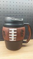 Front view of large Bubba Keg mug designed like a football with textured brown aesthetics and white laces detail, black base, and handle.
