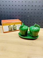 Green pepper-shaped salt and pepper shakers with matching tray in front of their box.