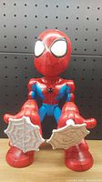 Front view of Spider-Man toy showing large head, red and blue costume, wide white eyes, web-shooting hands with white web attachments.