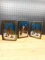 Three stained glass Christmas decoration panels displayed on a shelf against pegboard, showing festive winter scenes with children, animals, and snowmen on blue backgrounds.