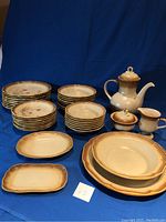 Overall view of the complete 38-piece Mikasa Whole Wheat dinnerware set with multiple stacks of plates, bowls, and serving pieces arranged on a blue cloth background.
