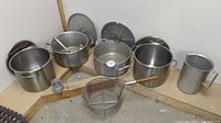 Photo showing large aluminum stock pots with matching lids, strainer basket, pitcher and metal utensils displayed on a wooden corner shelf.