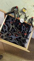 Wooden drawer filled with black plastic clamps with red tips, tape measure showing drawer size for scale.