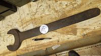 Full length and profile view of the large wrench on a workbench next to a pen for size comparison, showing overall shape and weathered condition.